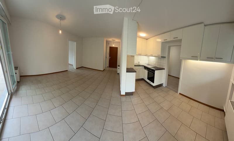 Empty kitchen and living area with tiled flooring, white cabinets, and built-in appliances, connected by an open doorway.
