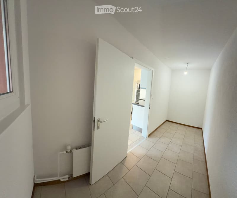 Empty hallway with tiled floor, white walls, and a radiator on the left side. An open door leads to another room with a visible kitchen area.