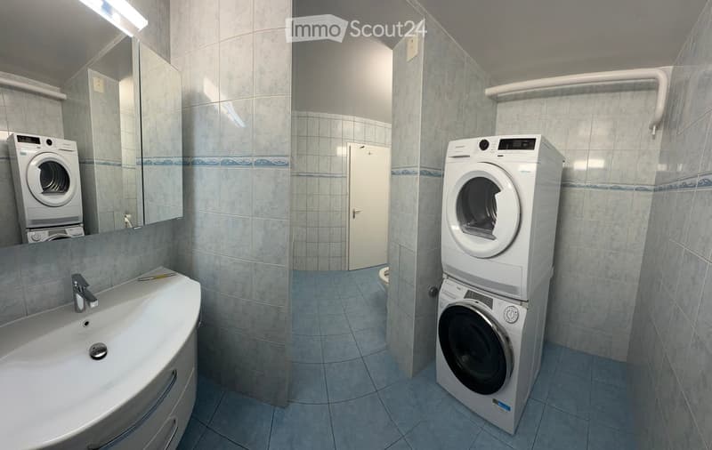 Modern bathroom with a sink, faucet, washing machine, dryer, and a large mirror. The room features tiled walls and a door leading to another area.