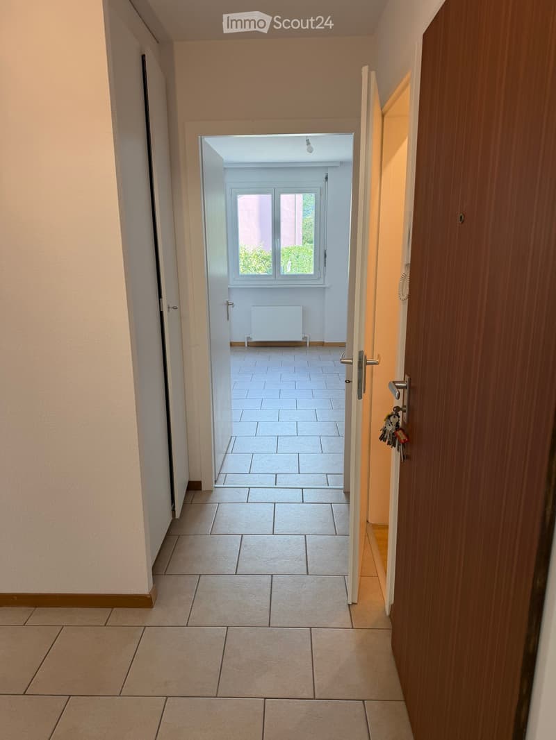 A clean, tiled hallway with beige walls and a wooden door on the right, leading to a bright room with large windows and a radiator.