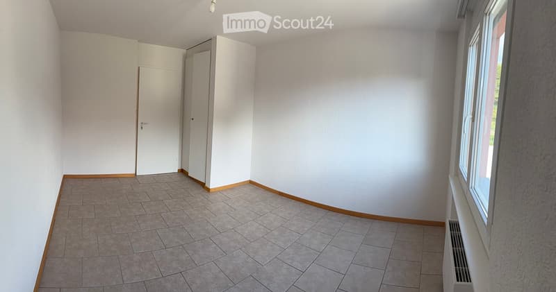 Empty room with tiled floor, white walls, and a window with a radiator below it. There are two white doors and a small cabinet in the background.