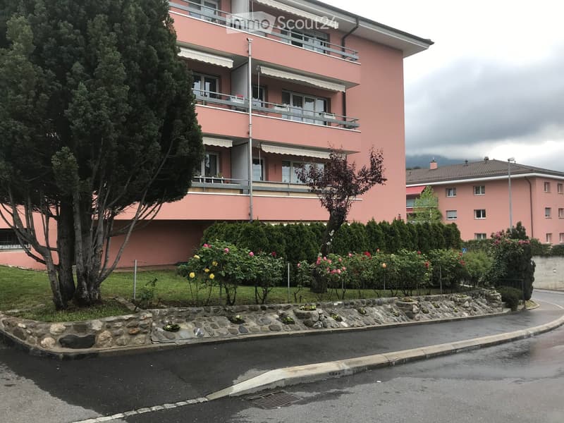 Pink multi-story apartment building with balconies, surrounded by greenery and flowers.