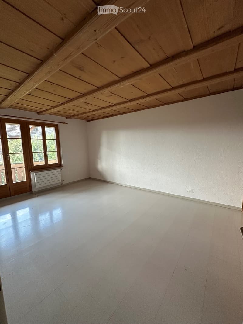 Empty room with white walls, polished concrete floor, wooden ceiling beams, and large windows with wooden frames.