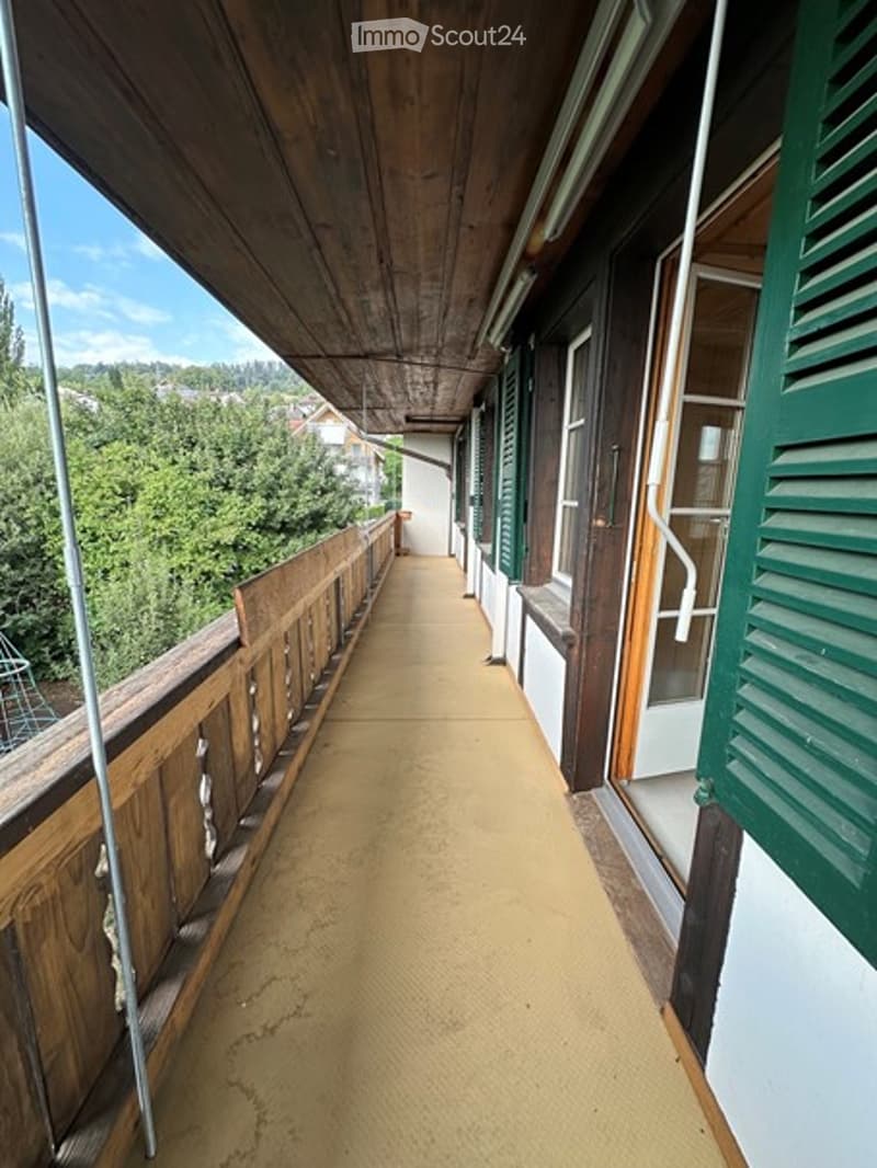Wooden balcony with green shutters, overlooking a green landscape with trees and houses.