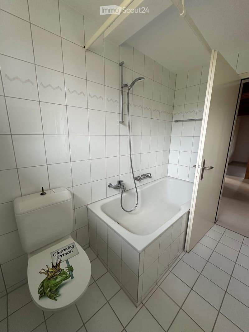 Bathroom with white tiled walls and floor, featuring a bathtub with a showerhead, a toilet with a decorative sticker, and a door leading to another room.