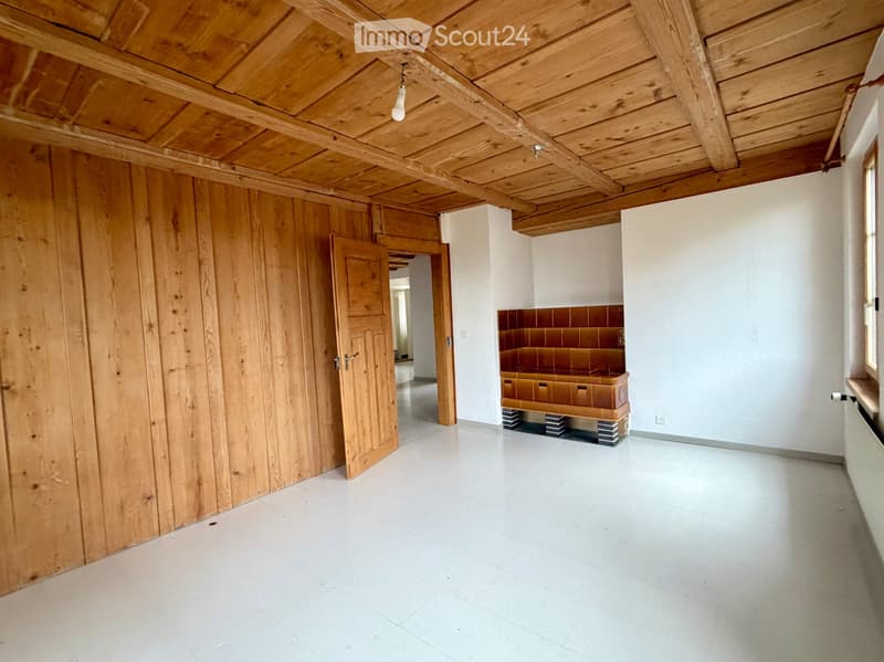 Empty room with wooden walls, exposed wooden ceiling beams, white tiled floor, and a small wooden bench with drawers in the corner.