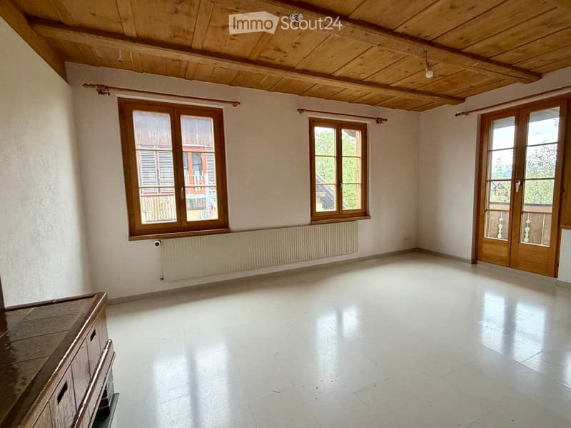 Empty room with white tiled floor, wooden ceiling beams, large windows with wooden frames, and a radiator under one window.
