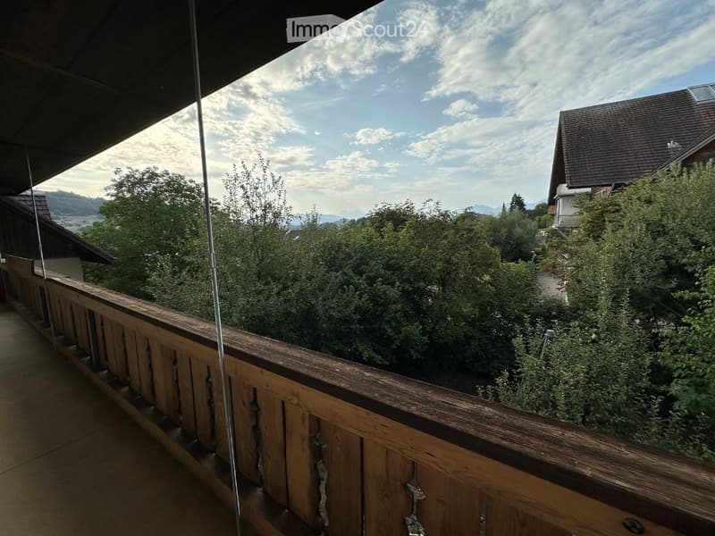 Balcony with wooden railing, overlooking a lush green garden and distant mountains under a partly cloudy sky.