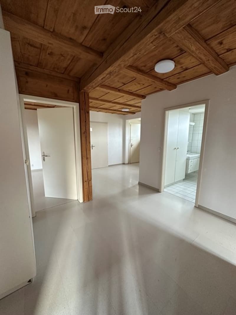 Empty room with wooden ceiling beams, white walls, and a smooth concrete floor. The space is completely unfurnished, with visible doorways leading to other rooms.
