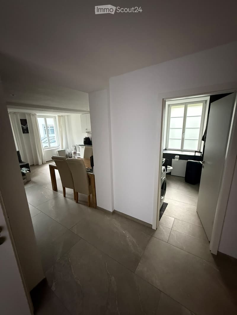 A modern, well-lit hallway leading to a dining area and a small bathroom. The dining area features a wooden table with four chairs and a laptop on the table. The bathroom includes a washing machine and a refrigerator.