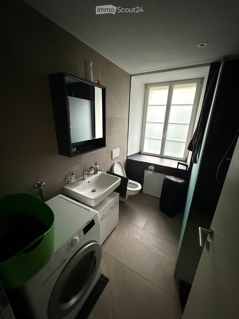 Modern bathroom with a sink, toilet, mirror cabinet, washing machine, and a window letting in natural light.