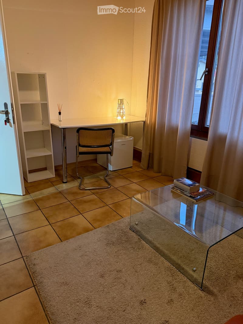 A furnished room with a tiled floor, a white desk with a chair, a glass coffee table with books, a shelf, a lamp, and a small refrigerator.