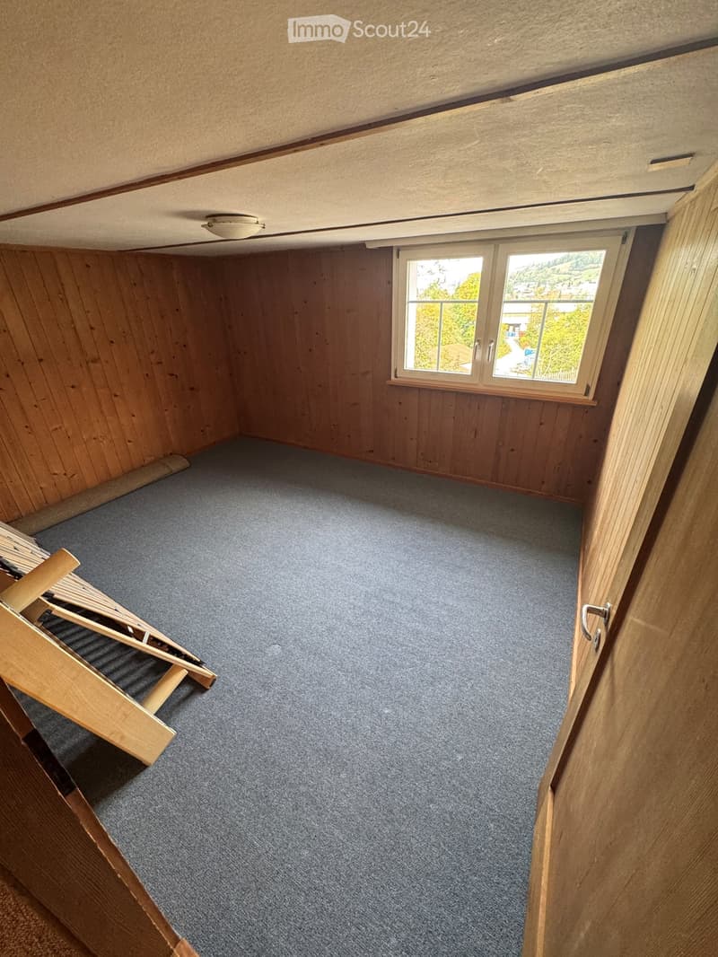 Completely empty room with wooden walls, carpeted floor, and a window with a view of the outdoors. No furniture or equipment is present.