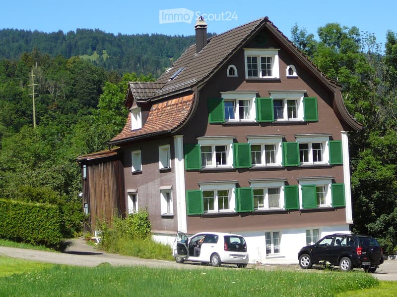 Two-story house with brown tiled roof, green shutters, and multiple windows. Two cars are parked in front of the house.
