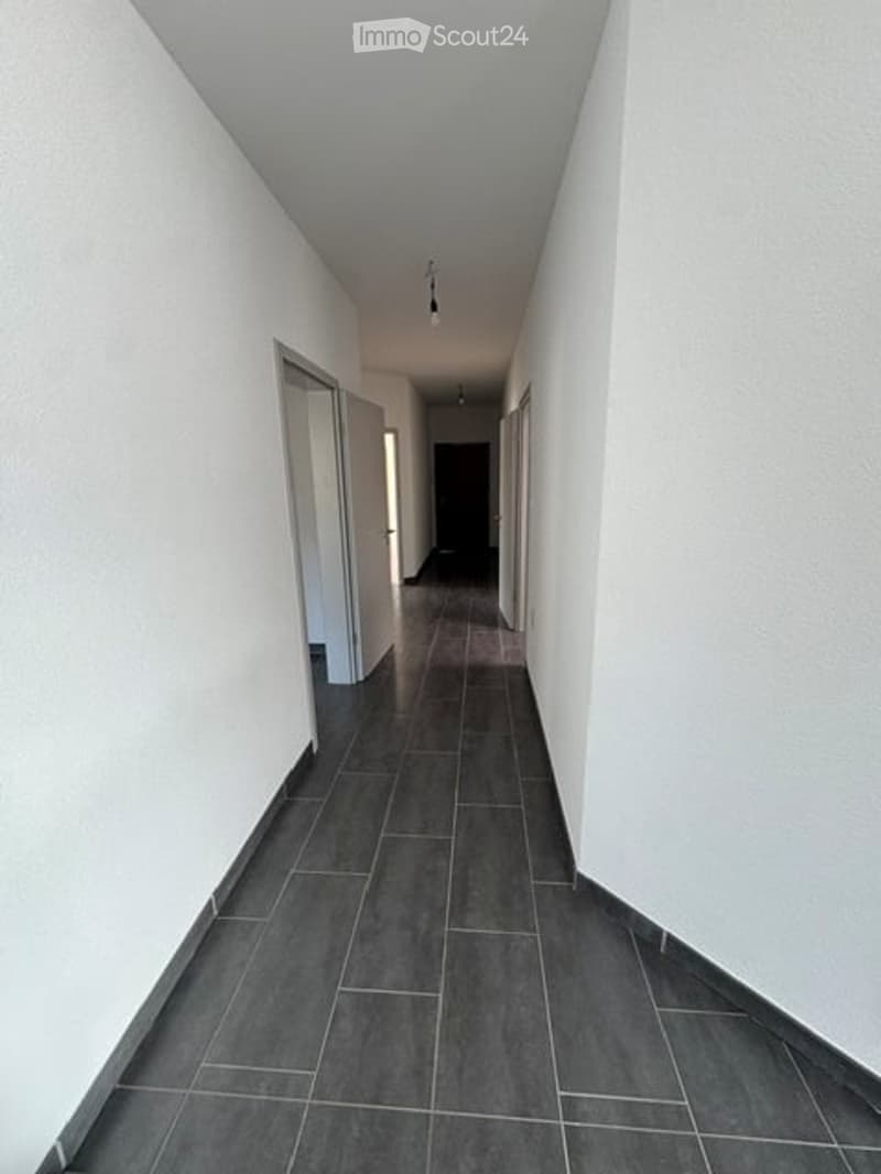 A long, narrow hallway with white walls, dark tiled flooring, and several doors on both sides. The ceiling has recessed lighting.