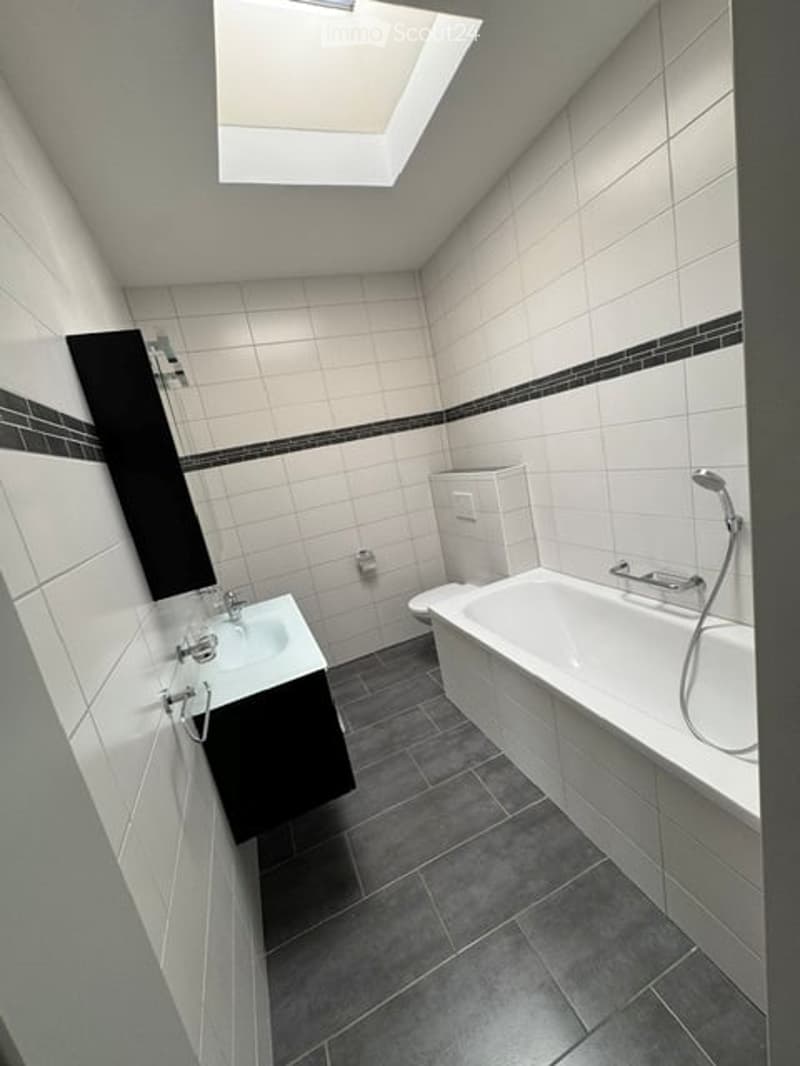 Modern bathroom with white tiles, dark grey floor, bathtub, sink, mirror, and toilet. Features a skylight window for natural light.