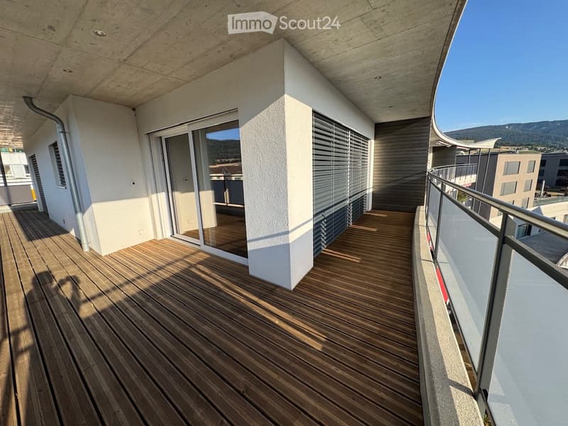 Wooden decked balcony with glass sliding doors, metal railing, and a view of the surrounding area.