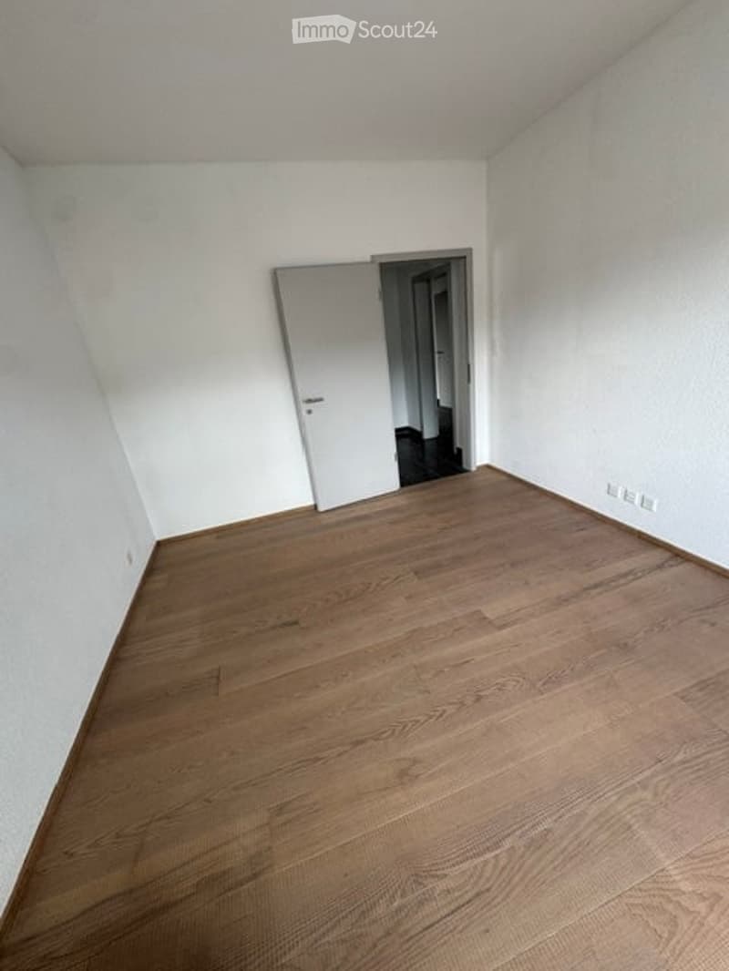 Empty room with light wooden flooring and white walls, featuring a closed white door leading to another space.