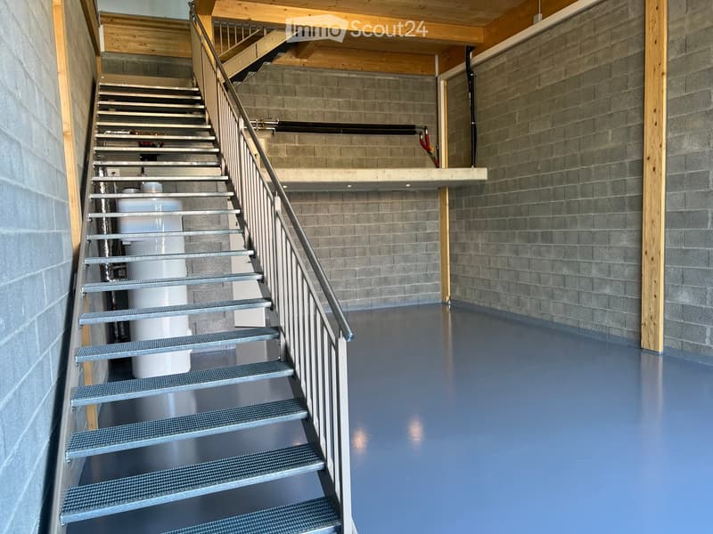 Modern staircase with metal railing and gray steps, leading to an upper level with exposed wooden beams and gray brick walls.