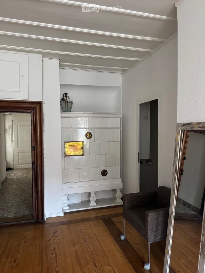 A furnished room with wooden flooring, a grey sofa, a white cabinet with shelves, and a decorative lamp on top.