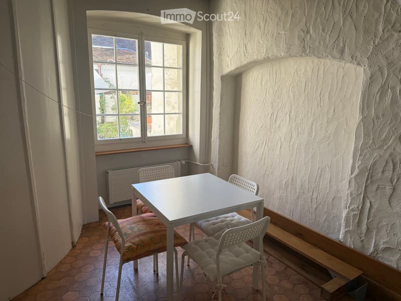 A small, furnished room with a white table and four chairs, a window with a view of the outside, and a radiator under the window.