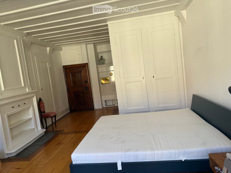Elegant bedroom with a large bed, wooden floor, white walls, and classic wooden doors. Features a small chair, a cabinet, and a nightstand.