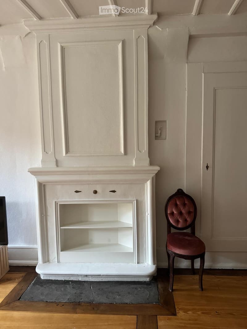 A room with a fireplace and a single chair, featuring wooden flooring and light-colored walls.