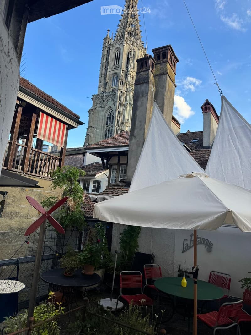 Courtyard with tables, chairs, umbrella, potted plants, and a view of a historic church tower.