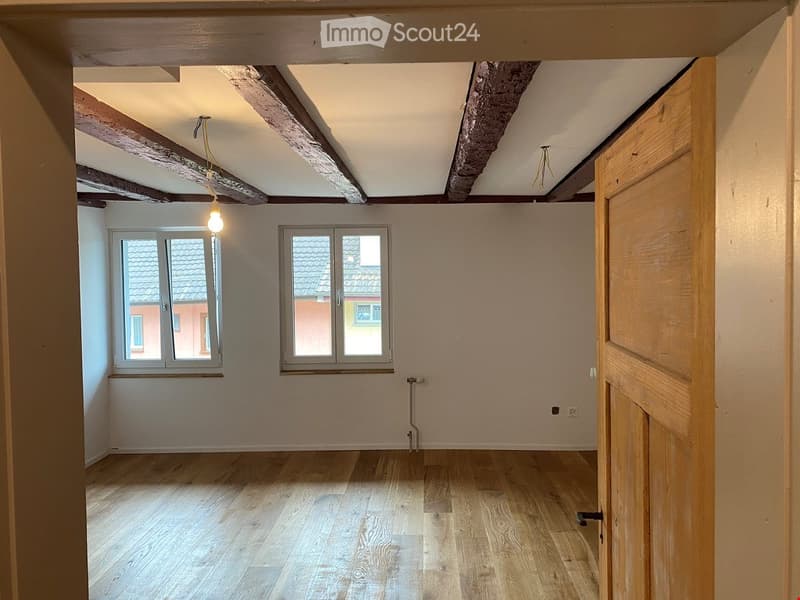 Empty room with wooden flooring, exposed wooden beams on the ceiling, two windows, and a single light fixture.