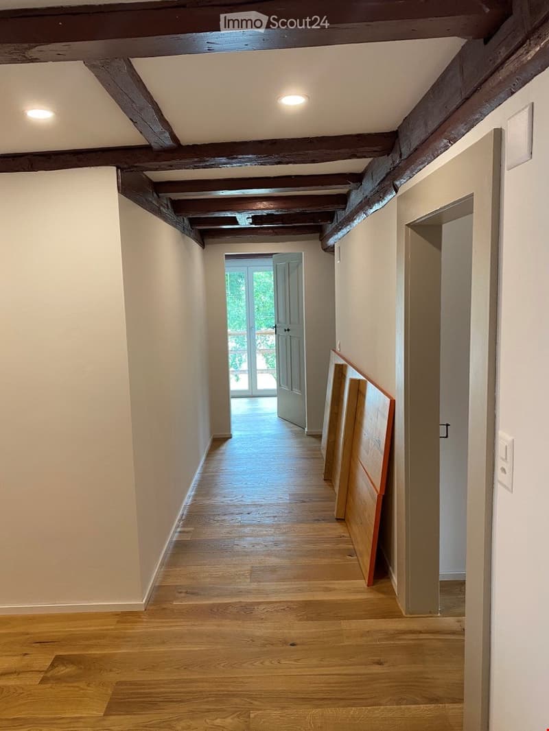A long, narrow hallway with wooden flooring and exposed wooden beams on the ceiling. The walls are painted a light neutral color, and there is a door at the end leading to a bright room with large windows.