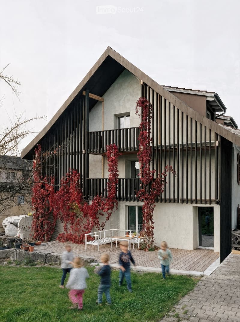 Modern two-story house with a unique geometric roofline, vertical wooden slats, and a balcony adorned with red climbing vines. The house features a wooden deck with outdoor furniture and a grassy lawn where children are playing.