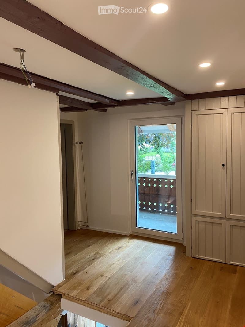 Empty room with wooden flooring, exposed wooden beams on the ceiling, and a sliding glass door leading to a balcony.