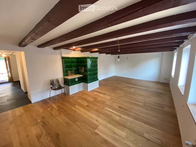 Empty room with wooden flooring, white walls, exposed wooden beams on the ceiling, and a small green tiled sink area in the corner.