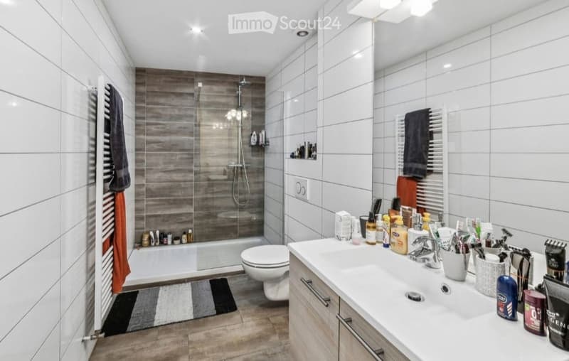 Modern bathroom with white tiles, a large sink with various toiletries, a toilet, a shower with a glass enclosure, and a towel rack with towels.