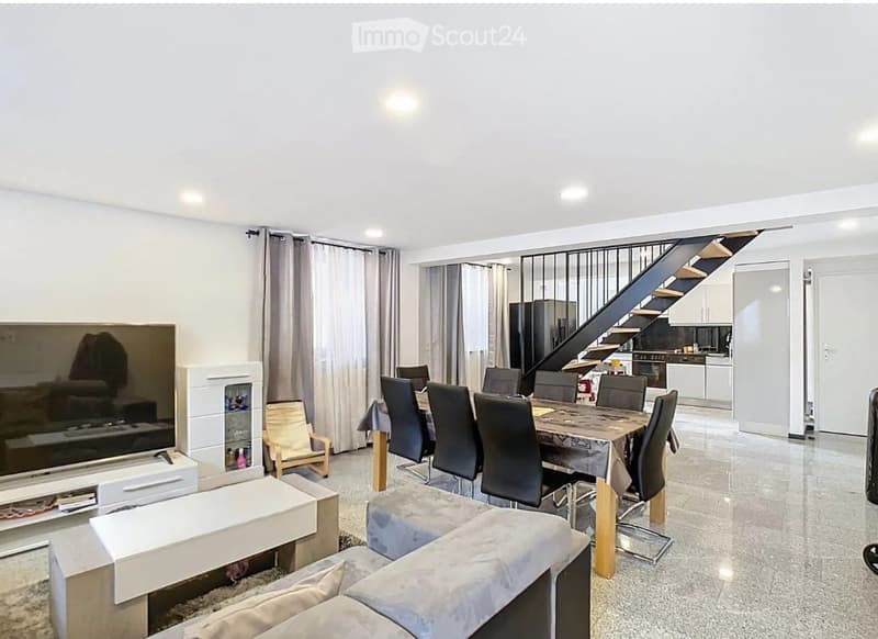 Open-plan living and dining area with a modern sofa, coffee table, dining table with chairs, and a television. A staircase leads to an upper level, and a kitchen area is visible in the background.