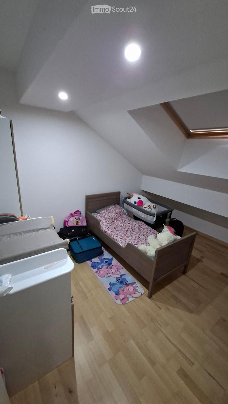 Cozy attic bedroom with wooden floor, slanted ceiling, single bed with pink bedding, crib, toys, and storage items.