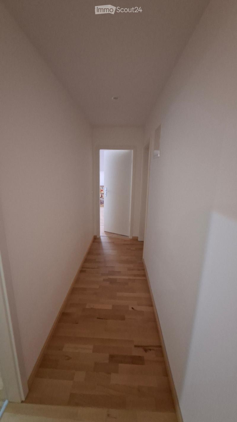 A narrow hallway with light wooden flooring and white walls, leading to an open doorway at the end.