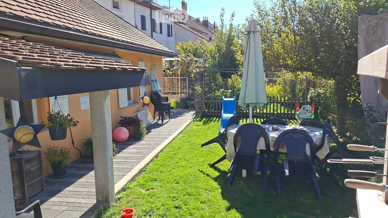 Garden with a dining table and chairs, a large umbrella, a small pathway, and a house with a balcony and potted plants.