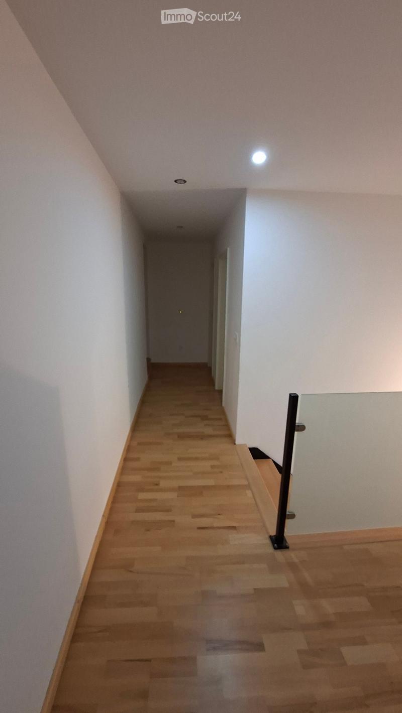 A narrow hallway with light wooden flooring and white walls, featuring a black handrail on the right side and recessed ceiling lighting.