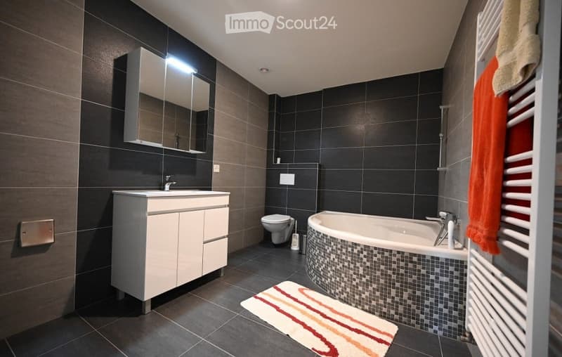 Modern bathroom with dark tiled walls and floor, white sink cabinet, mirror, toilet, bathtub, towel rack, and a colorful bath mat.