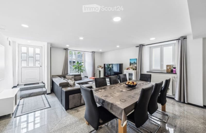 Open-plan living and dining area with a grey sectional sofa, coffee table, dining table with chairs, television, fireplace, and decorative plant.