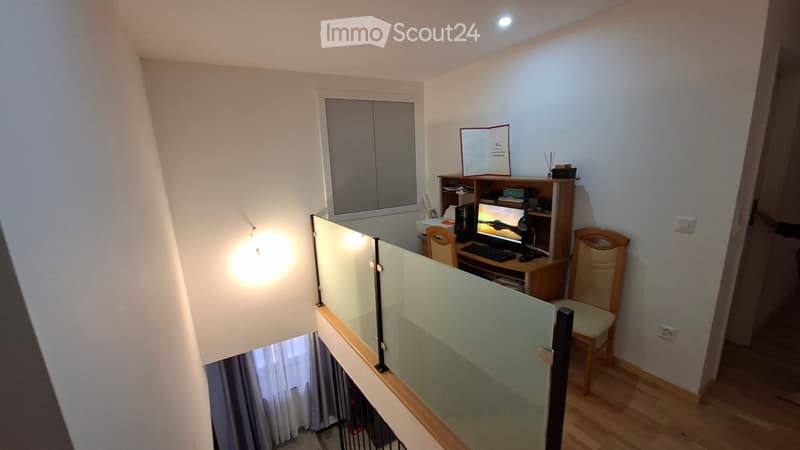 A modern, well-lit room with a desk setup, a computer, a chair, a lamp, and a cabinet. The room features a glass railing and wooden flooring.