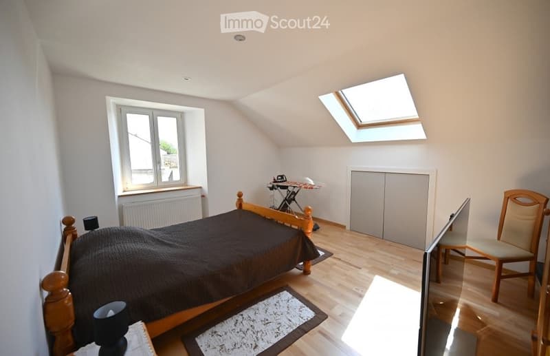 Cozy attic bedroom with wooden bed, chair, lamp, and built-in cabinet. Skylight window and radiator visible.