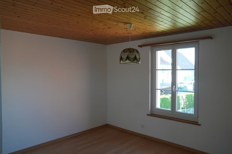 Empty room with wooden flooring, light grey walls, and a large window with a wooden sill. A hanging lamp is visible, and the ceiling is made of wood.