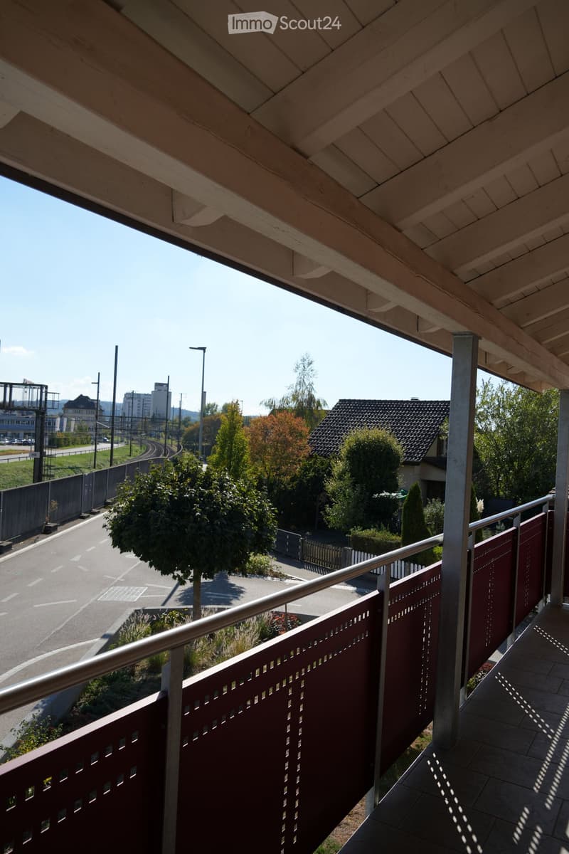 Balcony with metal railing, wooden decking, and view of street, trees, and buildings.