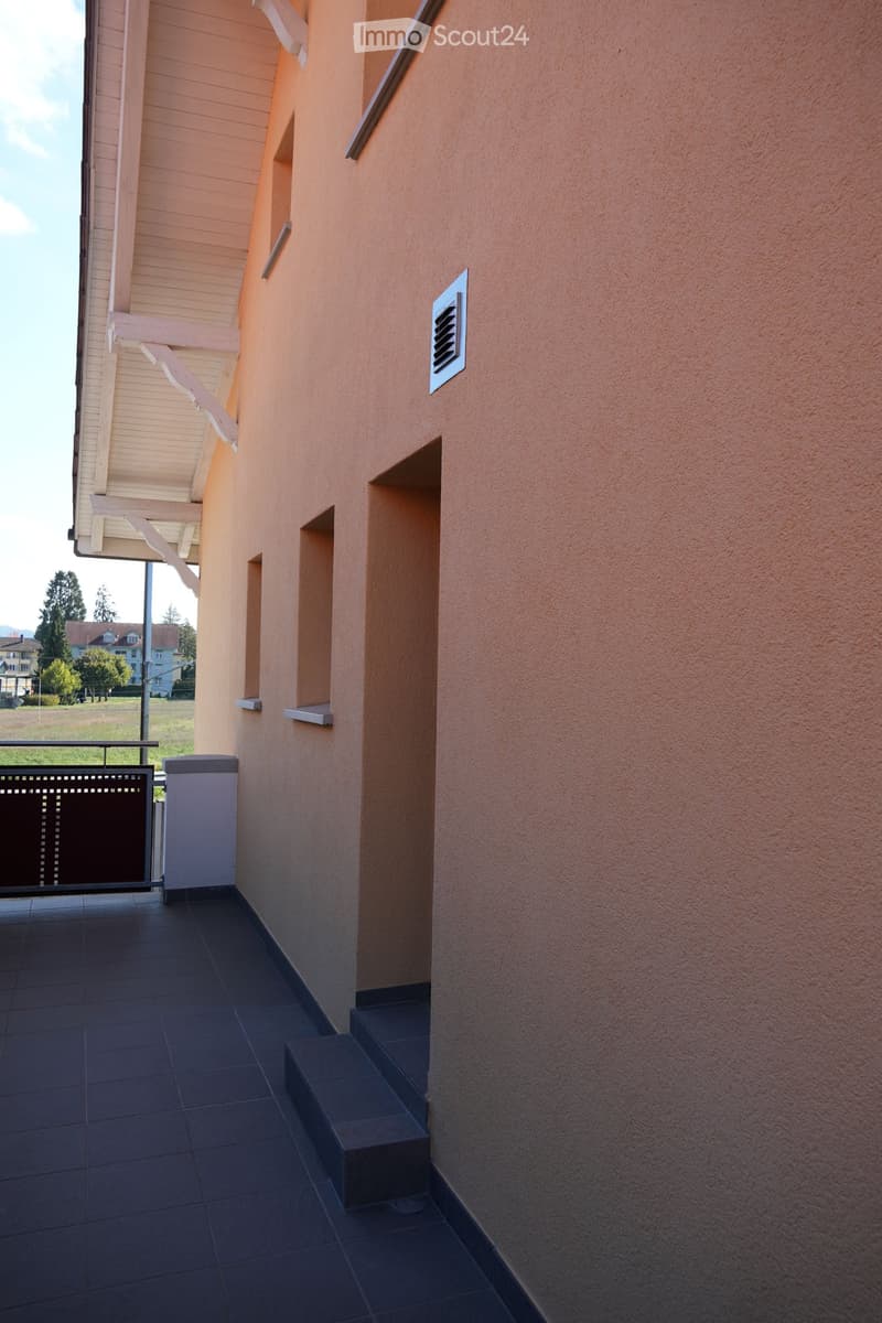 Balcony with dark tiled floor, metal railing, and steps leading up to a building with light-colored walls and a wooden overhang.