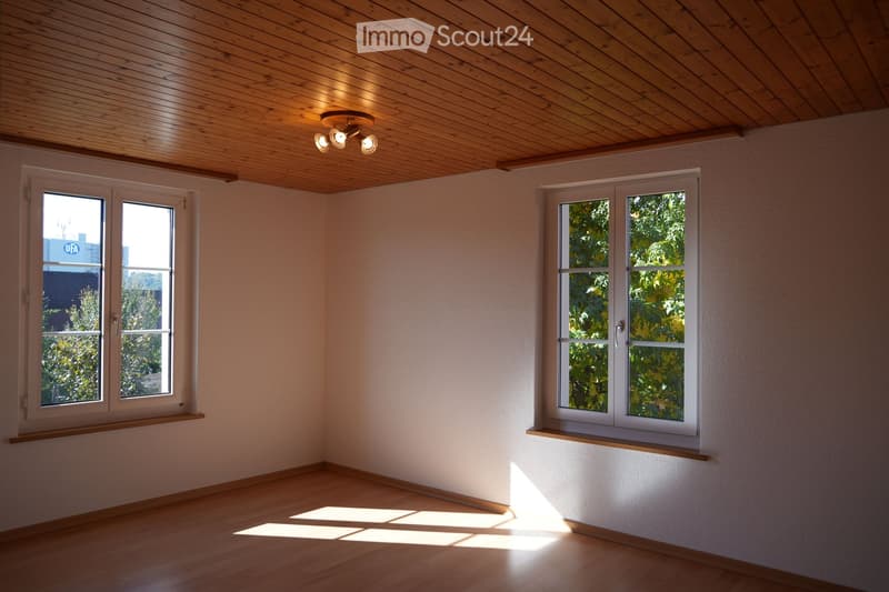 Empty room with wooden flooring, light-colored walls, and wooden ceiling. Two windows with white frames and wooden sills, one with a view of trees, the other showing a building with a 'UFA' sign. A ceiling light fixture is visible.