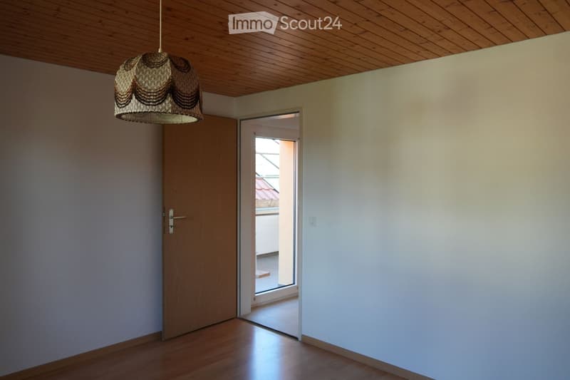Empty room with wooden flooring, white walls, a wooden door, and a hanging woven lamp. An open doorway leads to a balcony area with a tiled floor and a view of a roof.