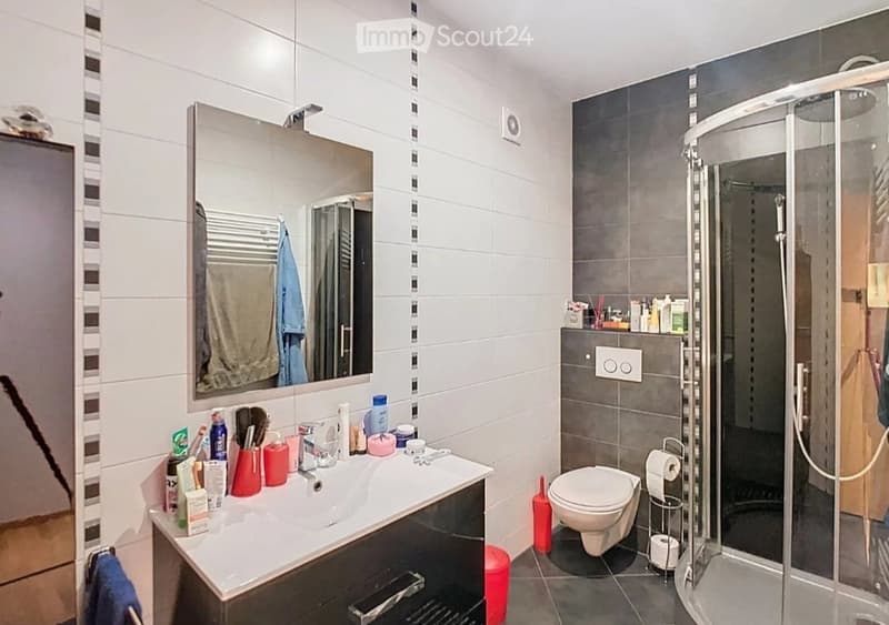 Modern bathroom with a white sink, large mirror, toilet, and glass-enclosed shower. The sink has various toiletries, and towels are neatly hung on a rack.