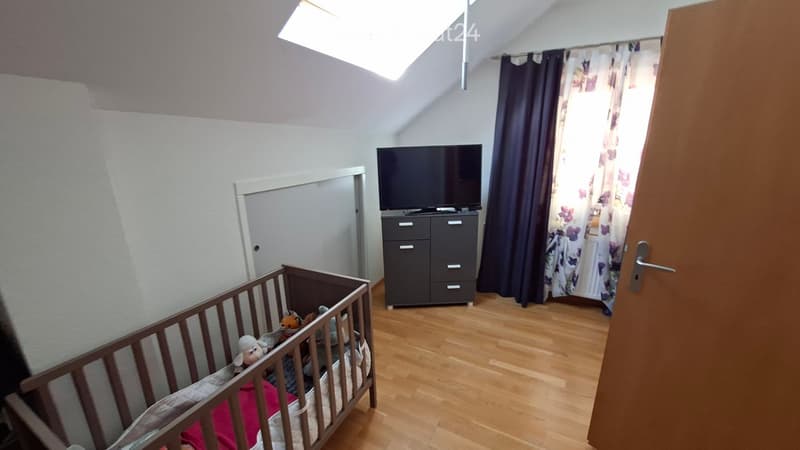 A small, well-lit room with wooden flooring, a crib, a dresser with a TV on top, and a window with curtains.